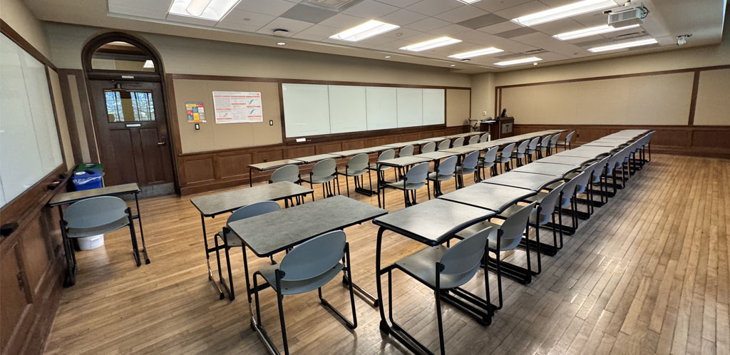 shows classroom with individual desk and chairs for students lined up in rows facing instructor podium in the background. Door to hallway is on same wall as podium.