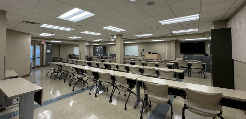 Brehm 130. Room includes long tables with chairs arranged around them. Instructor podium includes the control panel, instructor screen, and a spot for laptop. Next to the podium is a table that includes the document camera. The document camera table can be raised and lowered by pressing a button on the front of the table.