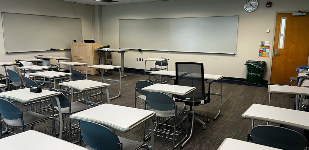 Bailey 425. Room includes singular desks with chairs. Instructor podium includes the control panel, instructor screen, and a spot for laptop. Next to the podium is a table that includes the document camera. The document camera table can be raised and lowered by pressing a button on the front of the table.