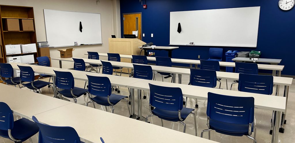 Bailey 428. Room includes long tables with chairs arranged around them. Instructor podium includes the control panel, instructor screen, and a spot for laptop. Next to the podium is a table that includes the document camera. The document camera table can be raised and lowered by pressing a button on the front of the table.