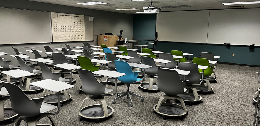 Buehler 334. Room includes node chairs that can be configured into different arrangements. Instructor podium includes the control panel, instructor screen, and a spot for laptop. Next to the podium is a table that includes the document camera. The document camera table can be raised and lowered by pressing a button on the front of the table.