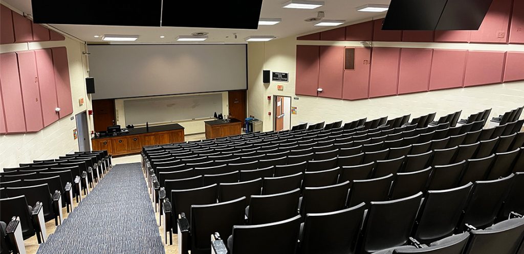 Buehler 555. Room includes theater style seating. Instructor podium includes the control panel, instructor screen, and a spot for laptop. Next to the podium is a table that includes the document camera. The document camera table can be raised and lowered by pressing a button on the front of the table.