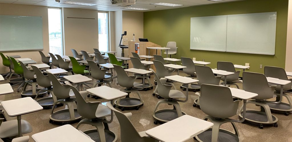 Burchfiel 101. Room includes node chairs that can be moved into different configurations. Instructor podium includes the control panel, instructor screen, and a spot for laptop. Next to the podium is a table that includes the document camera. The document camera table can be raised and lowered by pressing a button on the front of the table.