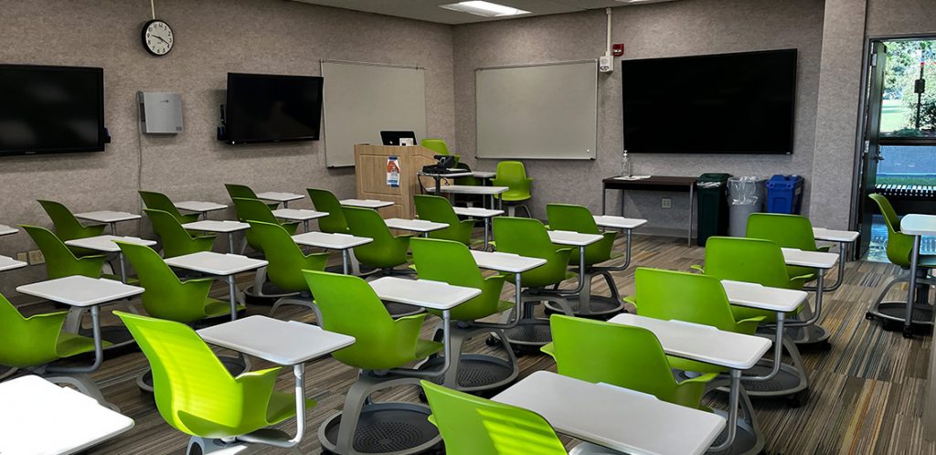 Communications 314. Room includes node chairs that can be moved into different configurations. Instructor podium includes the control panel, instructor screen, and a spot for laptop. Next to the podium is a table that includes the document camera. The document camera table can be raised and lowered by pressing a button on the front of the table.
