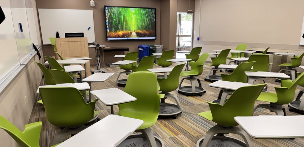 Communications room 314 showing instructor podium with wall monitor next to the podium and the door at the front of the room and students node chairs in the rest of the room.