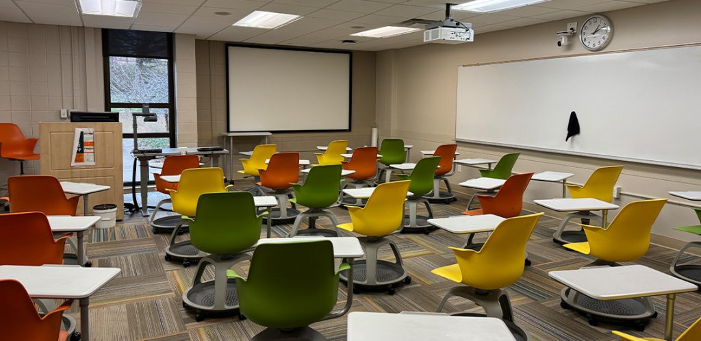 Humanities 53A. Room includes node chairs that can be moved into different configurations. Instructor podium includes the control panel, instructor screen, and a spot for laptop. Next to the podium is a table that includes the document camera. The document camera table can be raised and lowered by pressing a button on the front of the table.