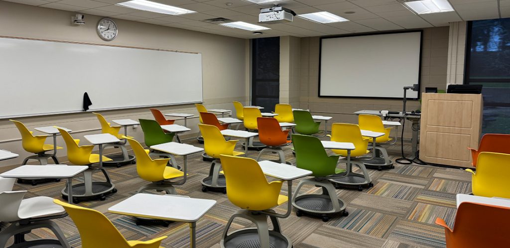 Humanities 56. Room includes node chairs that can be moved into different configurations. Instructor podium includes the control panel, instructor screen, and a spot for laptop. Next to the podium is a table that includes the document camera. The document camera table can be raised and lowered by pressing a button on the front of the table.