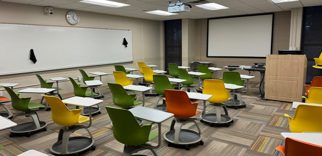 Humanities 57. Room includes node chairs that can be moved into different configurations. Instructor podium includes the control panel, instructor screen, and a spot for laptop. Next to the podium is a table that includes the document camera. The document camera table can be raised and lowered by pressing a button on the front of the table.