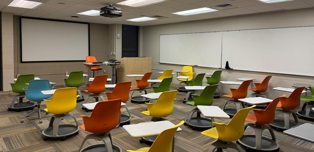 Humanities 59. Room includes node chairs that can be moved into different configurations. Instructor podium includes the control panel, instructor screen, and a spot for laptop. Next to the podium is a table that includes the document camera. The document camera table can be raised and lowered by pressing a button on the front of the table.