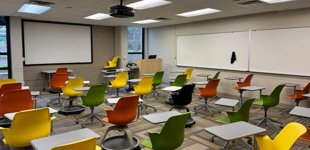 Humanities 60. Room includes node chairs that can be moved into different configurations. Instructor podium includes the control panel, instructor screen, and a spot for laptop. Next to the podium is a table that includes the document camera. The document camera table can be raised and lowered by pressing a button on the front of the table.