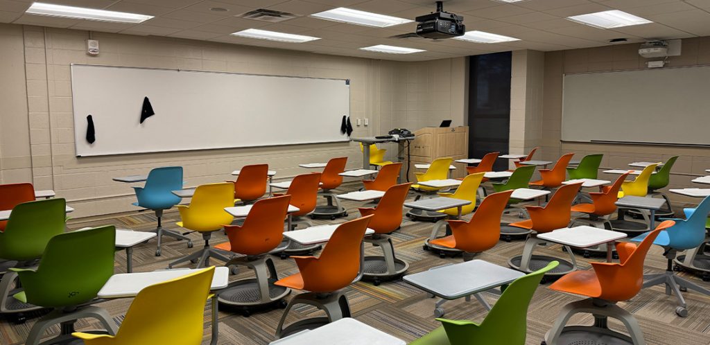 Humanities 61. Room includes node chairs that can be moved into different configurations. Instructor podium includes the control panel, instructor screen, and a spot for laptop. Next to the podium is a table that includes the document camera. The document camera table can be raised and lowered by pressing a button on the front of the table.