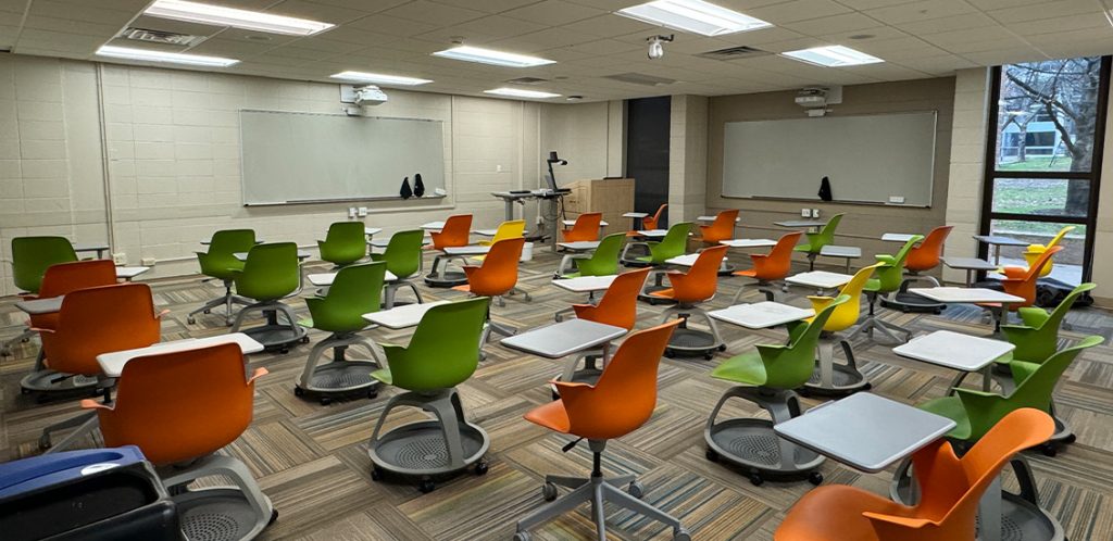 Humanities 63. Room includes node chairs that can be moved into different configurations. Instructor podium includes the control panel, instructor screen, and a spot for laptop. Next to the podium is a table that includes the document camera. The document camera table can be raised and lowered by pressing a button on the front of the table.
