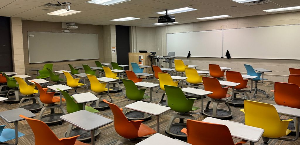 Humanities 64. Room includes node chairs that can be moved into different configurations. Instructor podium includes the control panel, instructor screen, and a spot for laptop. Next to the podium is a table that includes the document camera. The document camera table can be raised and lowered by pressing a button on the front of the table.