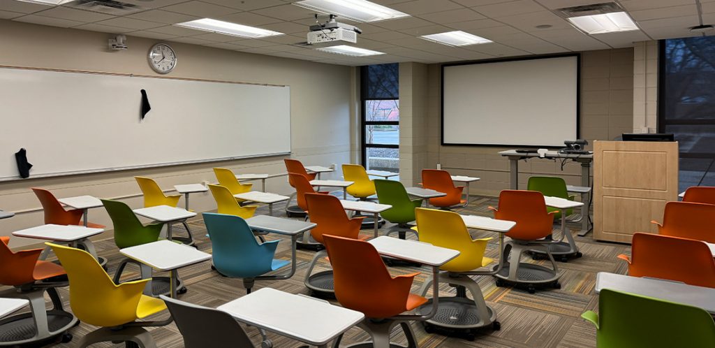 Humanities 69. Room includes node chairs that can be moved into different configurations. Instructor podium includes the control panel, instructor screen, and a spot for laptop. Next to the podium is a table that includes the document camera. The document camera table can be raised and lowered by pressing a button on the front of the table.