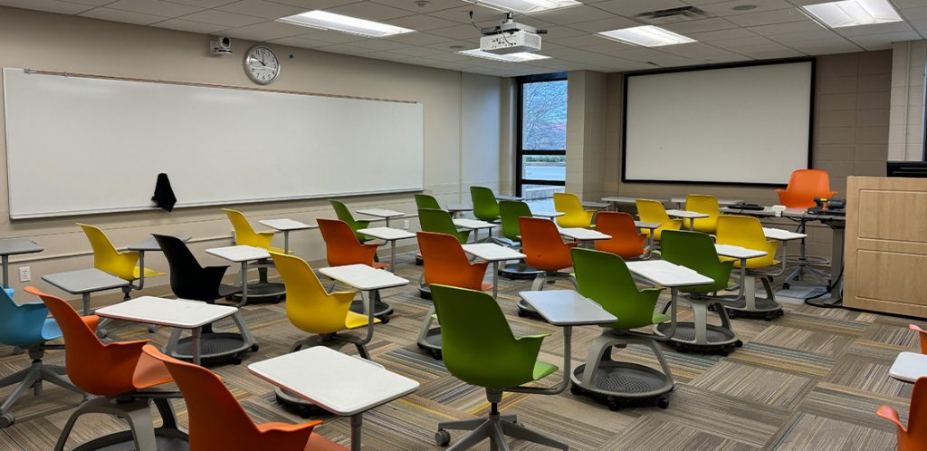 Humanities 70. Room includes node chairs that can be moved into different configurations. Instructor podium includes the control panel, instructor screen, and a spot for laptop. Next to the podium is a table that includes the document camera. The document camera table can be raised and lowered by pressing a button on the front of the table.