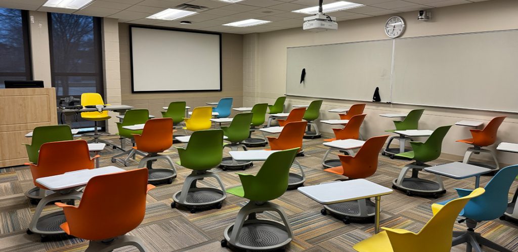 Humanities 71B. Room includes node chairs that can be moved into different configurations. Instructor podium includes the control panel, instructor screen, and a spot for laptop. Next to the podium is a table that includes the document camera. The document camera table can be raised and lowered by pressing a button on the front of the table.
