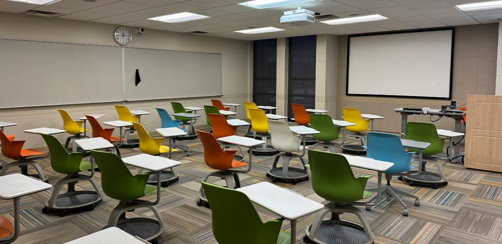 Humanities 71C. Room includes node chairs that can be moved into different configurations. Instructor podium includes the control panel, instructor screen, and a spot for laptop. Next to the podium is a table that includes the document camera. The document camera table can be raised and lowered by pressing a button on the front of the table.