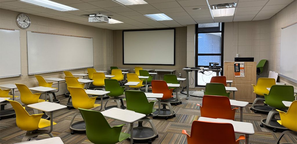 Humanities 101. Room includes node chairs that can be moved into different configurations. Instructor podium includes the control panel, instructor screen, and a spot for laptop. Next to the podium is a table that includes the document camera. The document camera table can be raised and lowered by pressing a button on the front of the table.