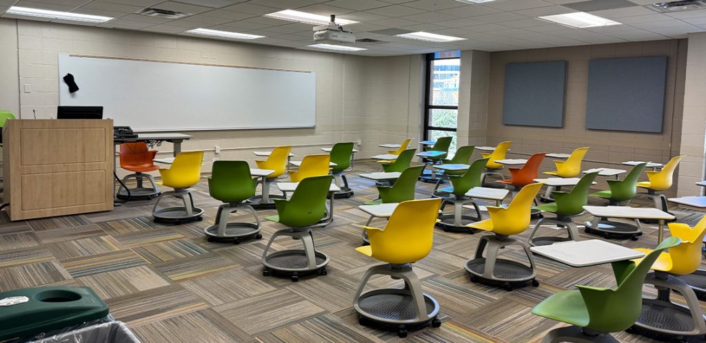 Humanities 102. Room includes node chairs that can be moved into different configurations. Instructor podium includes the control panel, instructor screen, and a spot for laptop. Next to the podium is a table that includes the document camera. The document camera table can be raised and lowered by pressing a button on the front of the table.