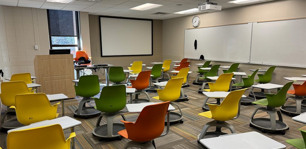 Humanities 103A. Room includes node chairs that can be moved into different configurations. Instructor podium includes the control panel, instructor screen, and a spot for laptop. Next to the podium is a table that includes the document camera. The document camera table can be raised and lowered by pressing a button on the front of the table.