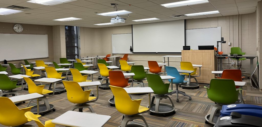 Humanities 103B. Room includes node chairs that can be moved into different configurations. Instructor podium includes the control panel, instructor screen, and a spot for laptop. Next to the podium is a table that includes the document camera. The document camera table can be raised and lowered by pressing a button on the front of the table.