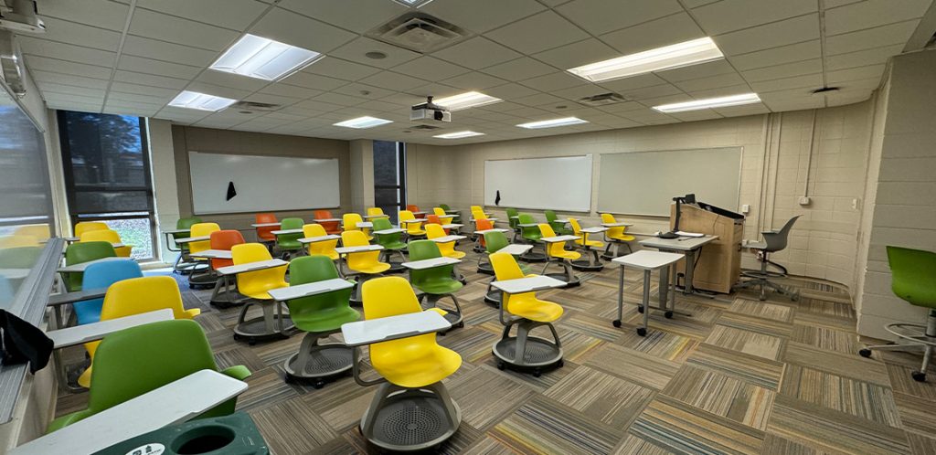 Humanities 104. Room includes node chairs that can be moved into different configurations. Instructor podium includes the control panel, instructor screen, and a spot for laptop. Next to the podium is a table that includes the document camera. The document camera table can be raised and lowered by pressing a button on the front of the table.