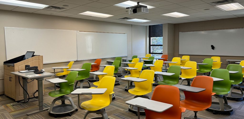 Humanities 105. Room includes node chairs that can be moved into different configurations. Instructor podium includes the control panel, instructor screen, and a spot for laptop. Next to the podium is a table that includes the document camera. The document camera table can be raised and lowered by pressing a button on the front of the table.