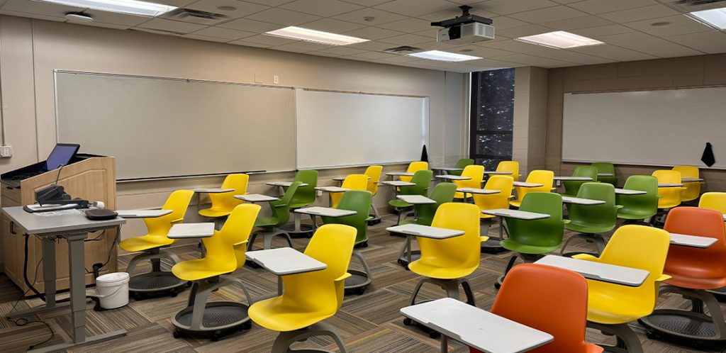 Humanities 106. Room includes node chairs that can be moved into different configurations. Instructor podium includes the control panel, instructor screen, and a spot for laptop. Next to the podium is a table that includes the document camera. The document camera table can be raised and lowered by pressing a button on the front of the table.