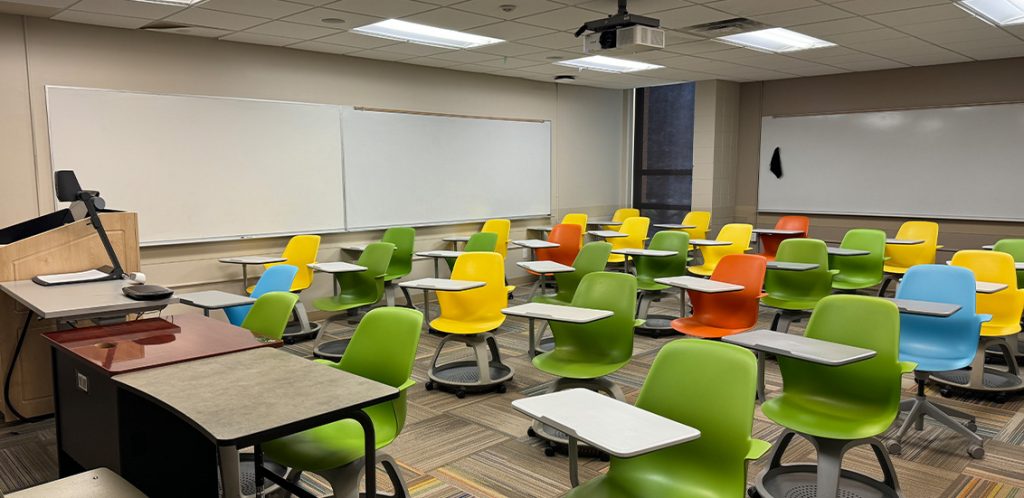 Humanities 107. Room includes node chairs that can be moved into different configurations. Instructor podium includes the control panel, instructor screen, and a spot for laptop. Next to the podium is a table that includes the document camera. The document camera table can be raised and lowered by pressing a button on the front of the table.