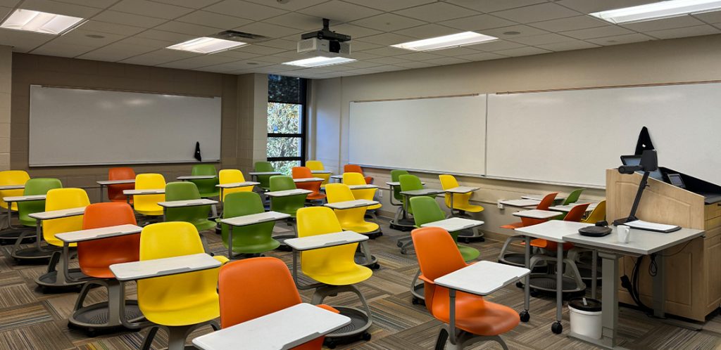 Humanities 108. Room includes node chairs that can be moved into different configurations. Instructor podium includes the control panel, instructor screen, and a spot for laptop. Next to the podium is a table that includes the document camera. The document camera table can be raised and lowered by pressing a button on the front of the table.