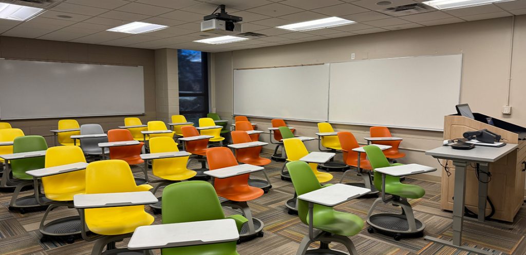 Humanities 109. Room includes node chairs that can be moved into different configurations. Instructor podium includes the control panel, instructor screen, and a spot for laptop. Next to the podium is a table that includes the document camera. The document camera table can be raised and lowered by pressing a button on the front of the table.