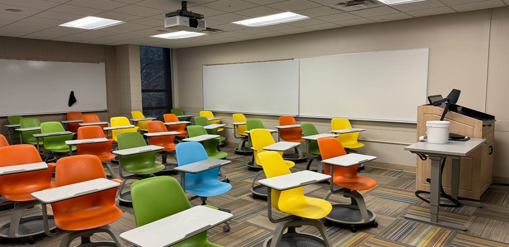 Humanities 110. Room includes node chairs that can be moved into different configurations. Instructor podium includes the control panel, instructor screen, and a spot for laptop. Next to the podium is a table that includes the document camera. The document camera table can be raised and lowered by pressing a button on the front of the table.