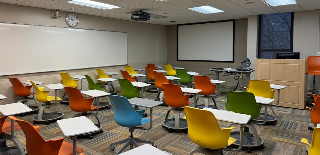 Humanities 112. Room includes node chairs that can be moved into different configurations. Instructor podium includes the control panel, instructor screen, and a spot for laptop. Next to the podium is a table that includes the document camera. The document camera table can be raised and lowered by pressing a button on the front of the table.