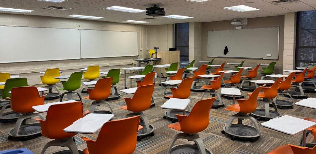 Humanities 113. Room includes node chairs that can be moved into different configurations. Instructor podium includes the control panel, instructor screen, and a spot for laptop. Next to the podium is a table that includes the document camera. The document camera table can be raised and lowered by pressing a button on the front of the table.