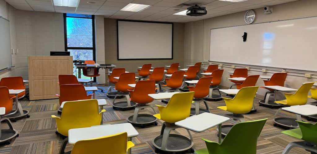 Humanities 115. Room includes node chairs that can be moved into different configurations. Instructor podium includes the control panel, instructor screen, and a spot for laptop. Next to the podium is a table that includes the document camera. The document camera table can be raised and lowered by pressing a button on the front of the table.