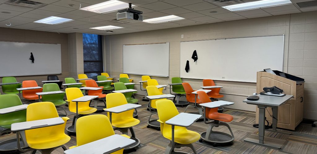 Humanities 118. Room includes node chairs that can be moved into different configurations. Instructor podium includes the control panel, instructor screen, and a spot for laptop. Next to the podium is a table that includes the document camera. The document camera table can be raised and lowered by pressing a button on the front of the table.