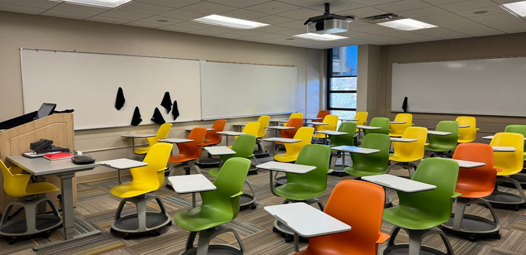 Humanities 120. Room includes node chairs that can be moved into different configurations. Instructor podium includes the control panel, instructor screen, and a spot for laptop. Next to the podium is a table that includes the document camera. The document camera table can be raised and lowered by pressing a button on the front of the table.