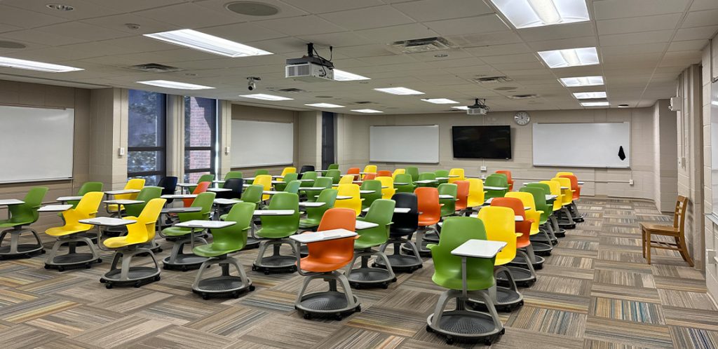 Humanities 203. Room includes node chairs that can be moved into different configurations. Instructor podium includes the control panel, instructor screen, and a spot for laptop. Next to the podium is a table that includes the document camera. The document camera table can be raised and lowered by pressing a button on the front of the table.