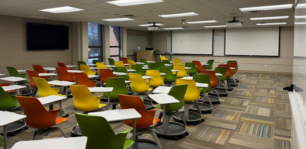 Humanities 205. Room includes node chairs that can be moved into different configurations. Instructor podium includes the control panel, instructor screen, and a spot for laptop. Next to the podium is a table that includes the document camera. The document camera table can be raised and lowered by pressing a button on the front of the table.