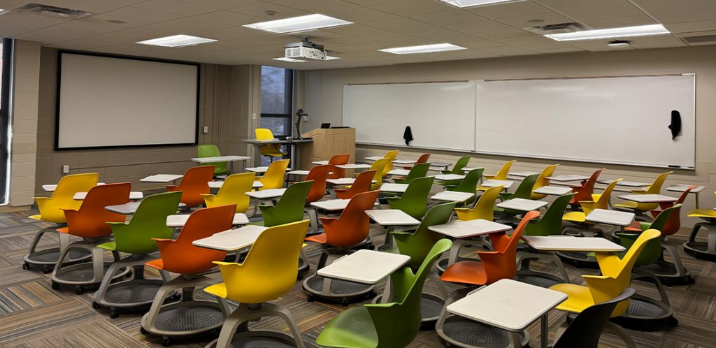 Humanities 205A. Room includes node chairs that can be moved into different configurations. Instructor podium includes the control panel, instructor screen, and a spot for laptop. Next to the podium is a table that includes the document camera. The document camera table can be raised and lowered by pressing a button on the front of the table.