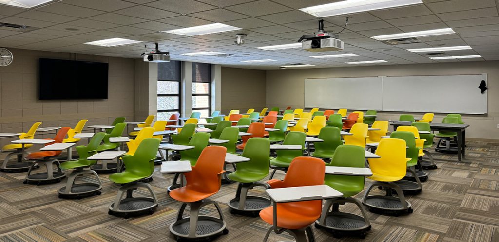 Humanities 207. Room includes node chairs that can be moved into different configurations. Instructor podium includes the control panel, instructor screen, and a spot for laptop. Next to the podium is a table that includes the document camera. The document camera table can be raised and lowered by pressing a button on the front of the table.