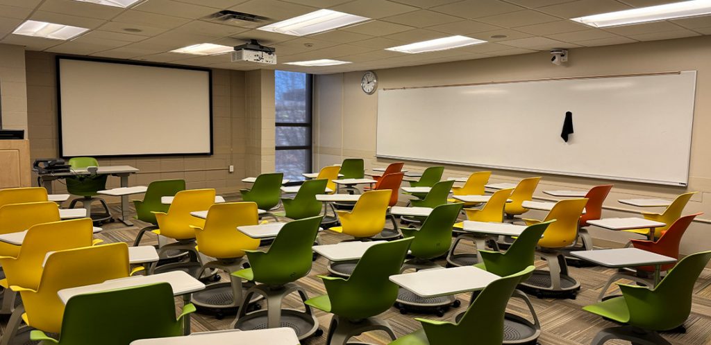 Humanities 215. Room includes node chairs that can be moved into different configurations. Instructor podium includes the control panel, instructor screen, and a spot for laptop. Next to the podium is a table that includes the document camera. The document camera table can be raised and lowered by pressing a button on the front of the table.