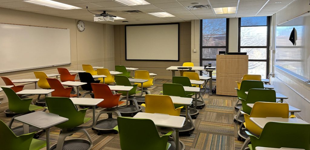 Humanities 217. Room includes node chairs that can be moved into different configurations. Instructor podium includes the control panel, instructor screen, and a spot for laptop. Next to the podium is a table that includes the document camera. The document camera table can be raised and lowered by pressing a button on the front of the table.