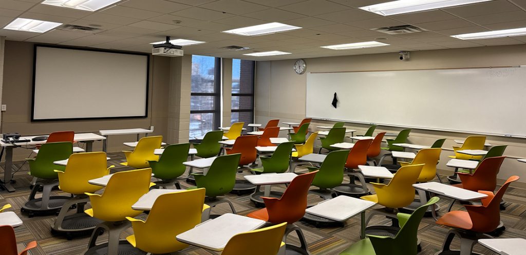 Humanities 218. Room includes node chairs that can be moved into different configurations. Instructor podium includes the control panel, instructor screen, and a spot for laptop. Next to the podium is a table that includes the document camera. The document camera table can be raised and lowered by pressing a button on the front of the table.