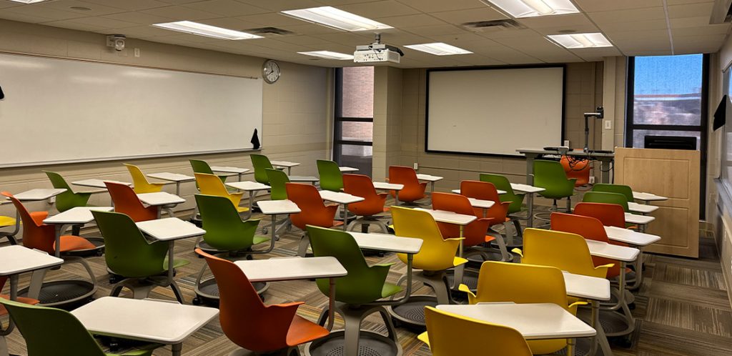 Humanities 220. Room includes node chairs that can be moved into different configurations. Instructor podium includes the control panel, instructor screen, and a spot for laptop. Next to the podium is a table that includes the document camera. The document camera table can be raised and lowered by pressing a button on the front of the table.