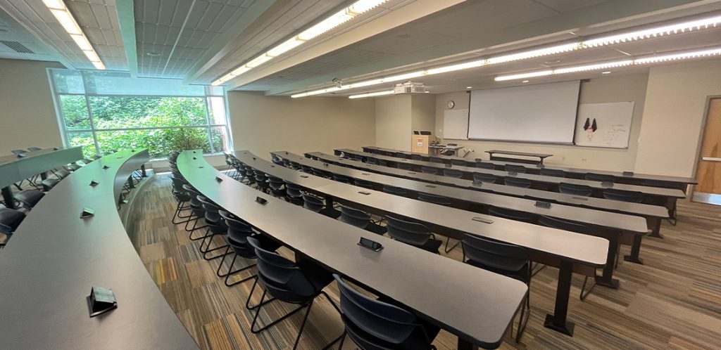 Min Kao 404. Room includes long tables and chairs. Instructor podium includes the control panel, instructor screen, and a spot for laptop. Next to the podium is a table that includes the document camera. The document camera table can be raised and lowered by pressing a button on the front of the table.