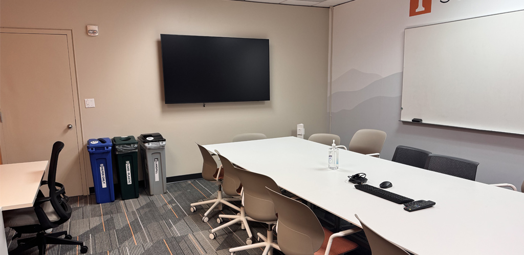 Stokely Management Center room 502 is set up like a conference room. Shows monitor on back wall, table with chairs in center and a table with chair to the left facing the wall.