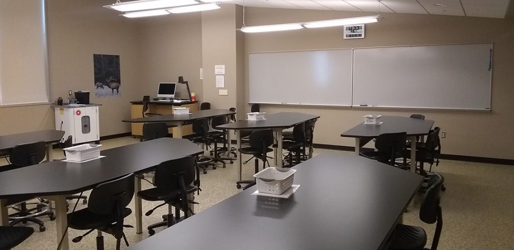 Strong 223. Room includes tables and chairs. Instructor podium includes the control panel, instructor screen, and a spot for laptop. Next to the podium is a table that includes the document camera.